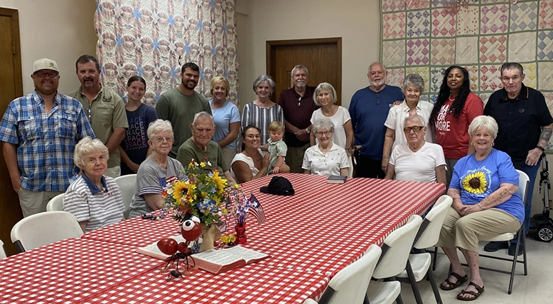 Group photo of participants and helpers at Trinity Fellowship stew for the soul event. Everyone is sitting/standing around a long table with red and white checker tableclothe and flowers in a vase