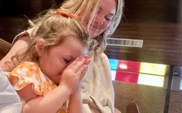 young girl with hands together held up to her face in prayer with her mother who has her arms around her